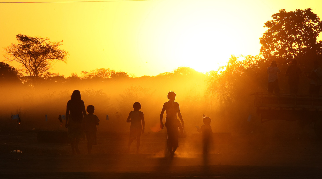 Silhuetas de várias pessoas, incluindo adultos e crianças, caminhando em direção a um pôr do sol brilhante. Preparativos para o Kauarup - Ritual Kuarup na aldeia Yawalapít, no Parque Indígena do Xingu (MT).