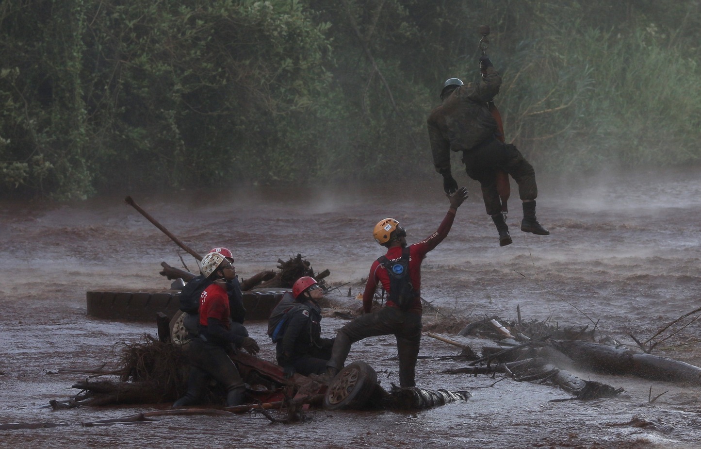 Trabalho de resgate após a tragédia de Brumadinho, com equipes de bombeiros e resgatistas em ação. Um dos profissionais é erguido por cordas, enquanto outros estão sobre o terreno afetado pela lama. A cena acontece em uma área com vegetação ao fundo, marcada pelos destroços da tragédia